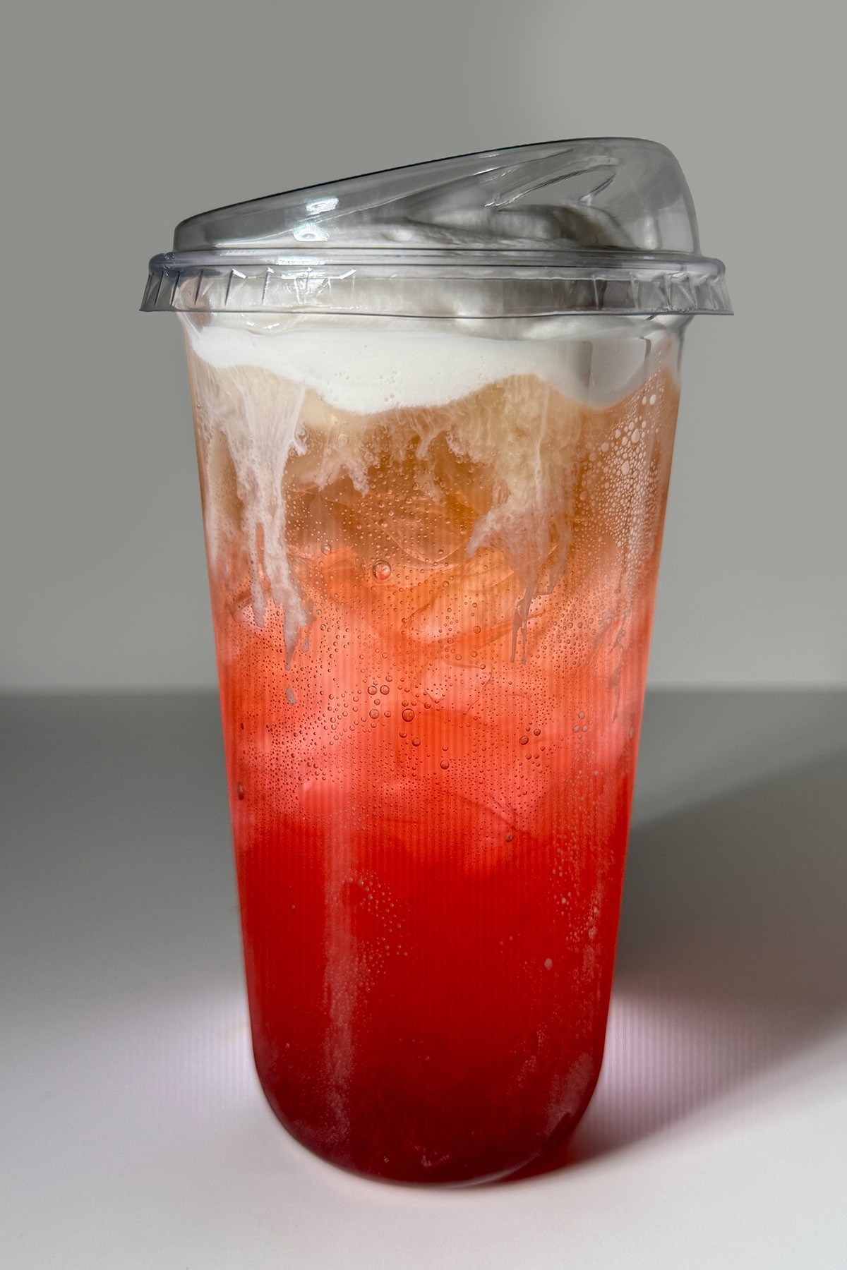 A striking, close-up photograph of a vibrant Desert Pear and Coconut Cream Soda in a clear, 16-ounce plastic cup against a minimalist white background. The drink features a dramatic visual gradient, starting with a deep, glowing magenta-red syrup at the base that transitions into a bubbly, translucent orange and golden-hue cream soda layer packed with ice. The carbonated layers are topped with a thick, velvety layer of white coconut cold foam that beautifully cascades down the inner walls of the cup. The beverage is sealed with a clear, slanted lid designed for sipping