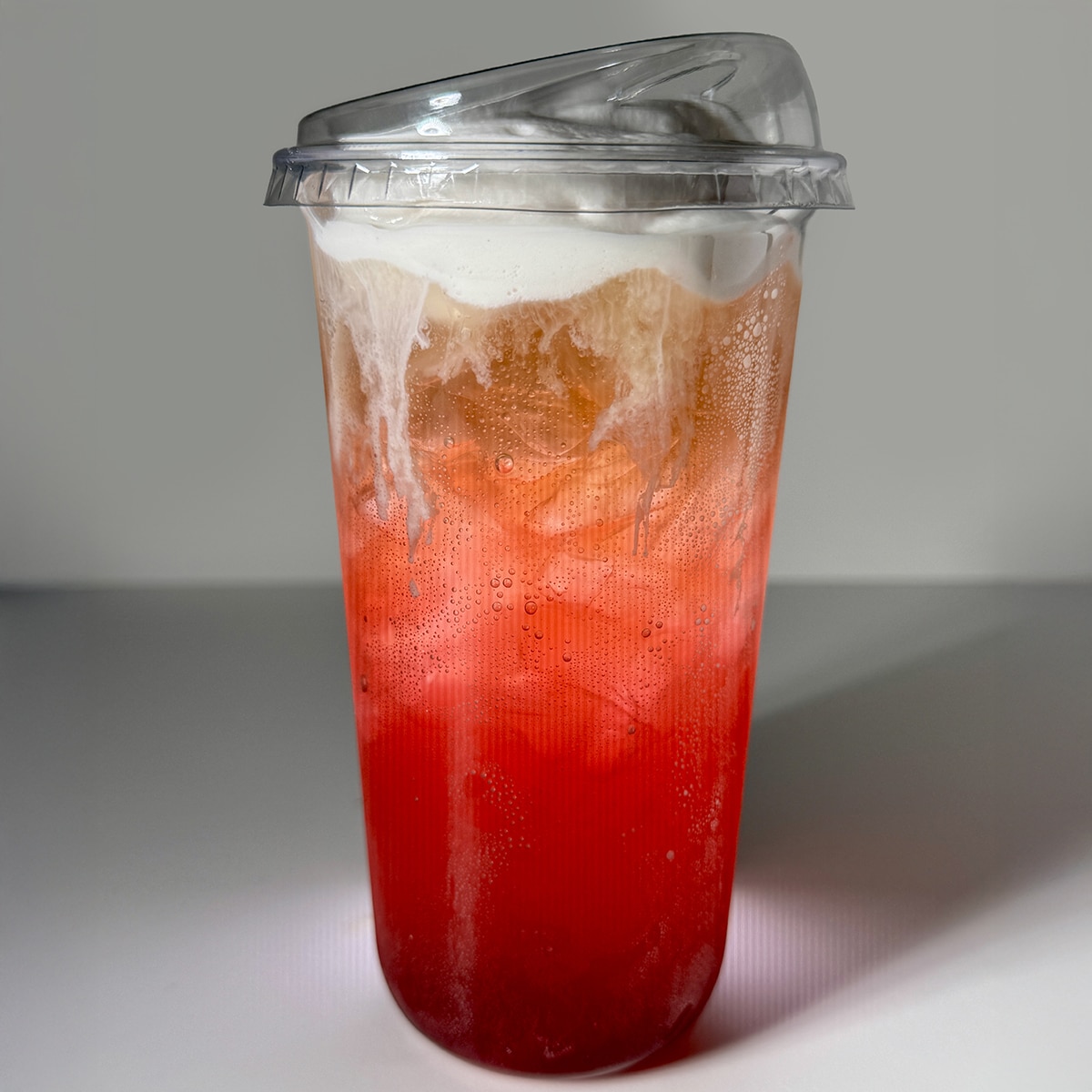 A striking, close-up photograph of a vibrant Desert Pear and Coconut Cream Soda in a clear, 16-ounce plastic cup against a minimalist white background. The drink features a dramatic visual gradient, starting with a deep, glowing magenta-red syrup at the base that transitions into a bubbly, translucent orange and golden-hue cream soda layer packed with ice. The carbonated layers are topped with a thick, velvety layer of white coconut cold foam that beautifully cascades down the inner walls of the cup. The beverage is sealed with a clear, slanted lid designed for sipping without a straw, captured with sharp, intentional lighting that highlights the condensation and effervescent bubbles on the plastic surface.