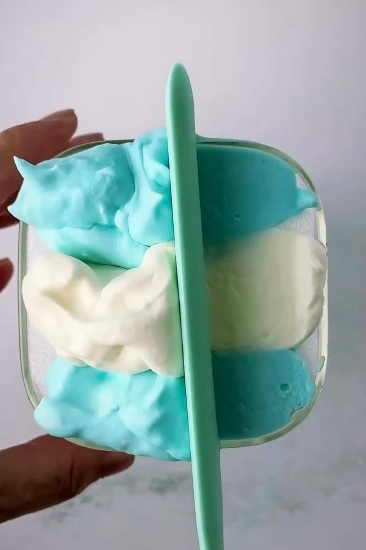 A real-life step-by-step tutorial shot showing a hand using a mint green spatula tool to level out the homemade cold foam stripes inside a square glass. Thick blocks of blue and white frothed heavy cream are being smoothed flush with the rim of the glass to create the clean, geometric horizontal stripes of the flag design.