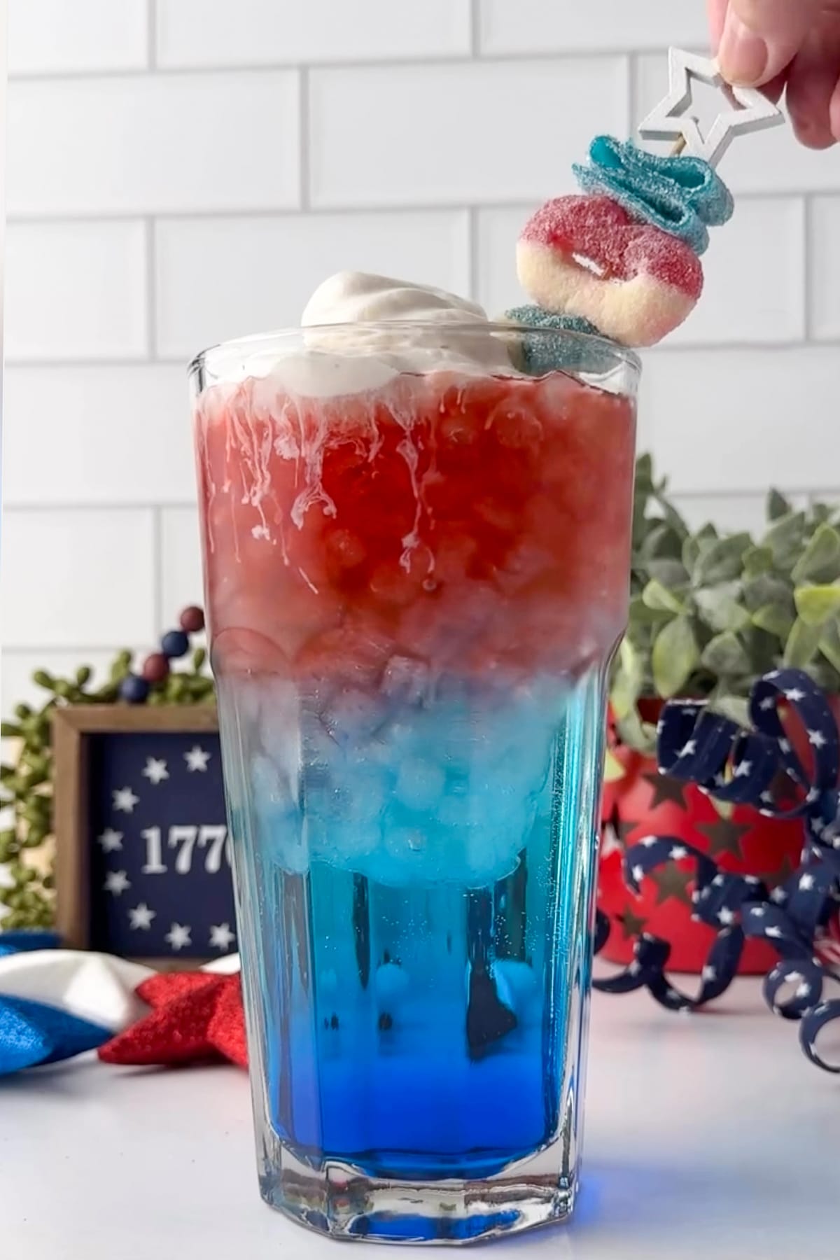 A close-up, master-level photograph of a completed Day 6 Patriotic Layered Dirty Soda in a tall glass cup. The drink features clear red, white, and blue layers. The dark blue syrup anchors the bottom, transitioning into a light blue soda middle, and a bright red strawberry top layer. Fluffy canned cold foam sits over the red surface, paired with a themed candy skewer garnish.