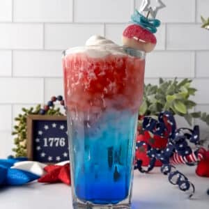 A close-up shot of a hand placing a themed candy skewer across the rim of a layered red, white, and blue dirty soda. The cocktail skewer features red and blue candy O's and a ribbon of sour candy strip beneath a silver star topper. The thick cold foam topping holds the skewer in place as the cream slowly melts down into the red soda layer.
