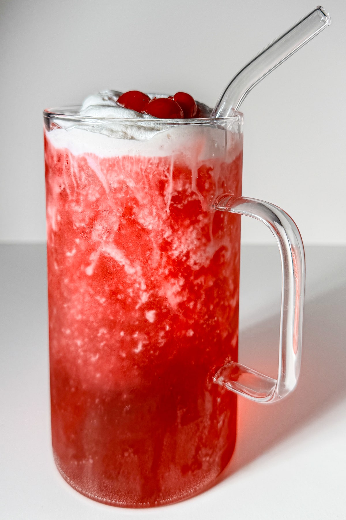 A bright red, bubbly Vanilla Shirley Temple dirty soda in a tall glass mug with a handle, filled with ice and topped with thick white canned cold foam and maraschino cherries.
