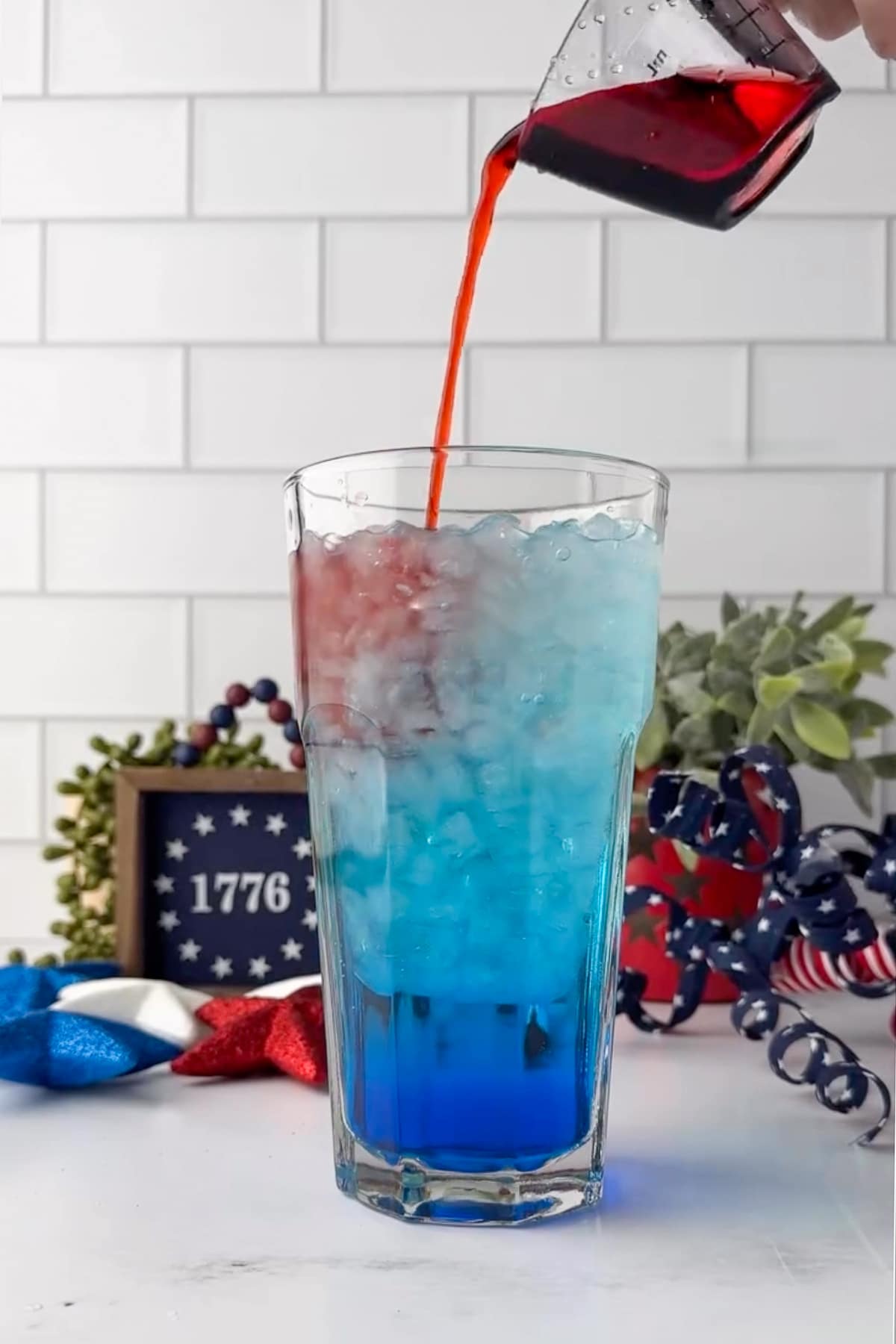 A real-life, action-focused tutorial shot of a hand pouring sugar-free strawberry syrup into a tall glass filled with ice. The red liquid hits the top of a bright blue soda base. The layers remain perfectly intact without bleeding together. The scene is set against a clean white subway tile backsplash with festive, patriotic decor blurred in the background.